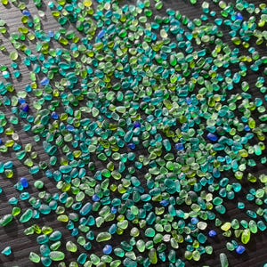 Op de afbeelding: Een close-up afbeelding van een grote verzameling kleine, kleurrijke glazen kralen. De kralen zijn voornamelijk groen en blauw, met wat heldere en gele kralen ertussen. De kralen zijn verspreid over een donker oppervlak.