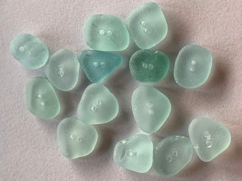 K&ouml;nnte beinhalten: Eine Sammlung von 14 hellblauen, glatten Muschelkn&ouml;pfen mit jeweils zwei L&ouml;chern. Die Kn&ouml;pfe haben eine nat&uuml;rliche, unregelm&auml;&szlig;ige Form.