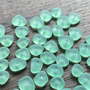 May include: A collection of light green sea glass beads, each with a hole in the centre. The beads are smooth and have a slightly irregular shape.