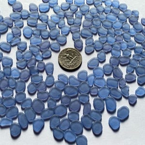 May include: A collection of small, rounded, blue sea glass pieces scattered on a white surface. A US quarter coin is in the center, providing a size reference. The sea glass has a frosted appearance and varying shades of blue.
