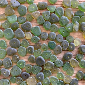 Puede incluir: Una colecci&oacute;n de cuentas de vidrio marino en varios tonos de verde y amarillo, cada una con un peque&ntilde;o agujero perforado en el centro. Las cuentas est&aacute;n esparcidas sobre una superficie de madera, mostrando sus formas irregulares y texturas esmeriladas.