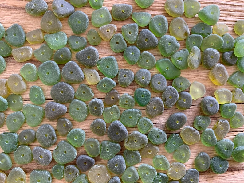Puede incluir: Una colecci&oacute;n de cuentas de vidrio marino en varios tonos de verde y amarillo, cada una con un peque&ntilde;o agujero perforado en el centro. Las cuentas est&aacute;n esparcidas sobre una superficie de madera, mostrando sus formas irregulares y texturas esmeriladas.