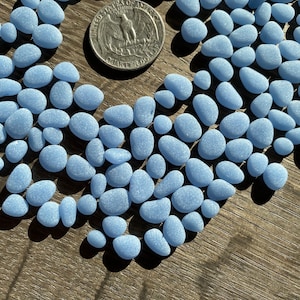May include: A large quantity of light blue glass pebbles, scattered on a wooden surface. The pebbles are smooth and have a frosted finish. A US quarter is included for scale.