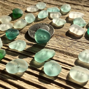 K&ouml;nnte beinhalten: Eine Sammlung von Meeresglas-Kn&ouml;pfen in Gr&uuml;n- und Wei&szlig;t&ouml;nen. Die Kn&ouml;pfe sind in einem Muster auf einer Holzoberfl&auml;che angeordnet. Eine US-amerikanische 25-Cent-M&uuml;nze dient als Gr&ouml;&szlig;envergleich.