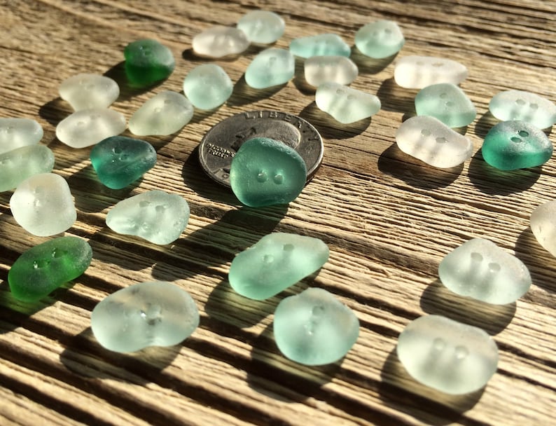 K&ouml;nnte beinhalten: Eine Sammlung von Meeresglas-Kn&ouml;pfen in Gr&uuml;n- und Wei&szlig;t&ouml;nen. Die Kn&ouml;pfe sind in einem Muster auf einer Holzoberfl&auml;che angeordnet. Eine US-amerikanische 25-Cent-M&uuml;nze dient als Gr&ouml;&szlig;envergleich.