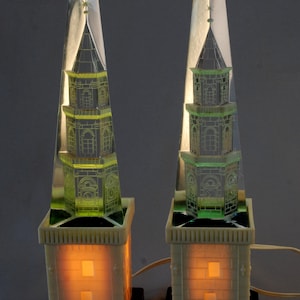Peut inclure: Deux lampes en verre transparent en forme de flèche avec une base verte et une base blanche avec une lumière à l'intérieur. La flèche est gravée d'un bâtiment détaillé.