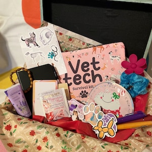 May include: A pink and white gift box filled with various items, including a pink pouch with the text "Vet tech survival kit", a pink and white plate with a cartoon dog, a blue hair scrunchie, a purple pen, an orange pen, a yellow balloon animal, a small pink and white dog toy, a lavender hand cream, a box of peppermint bark tea, and a wooden beaded bracelet.