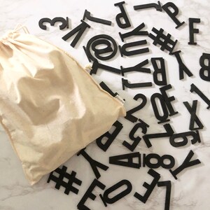 May include: A white canvas bag with black letters, numbers, and symbols scattered around it. The letters include the alphabet, numbers, and symbols like the at symbol and the pound sign.