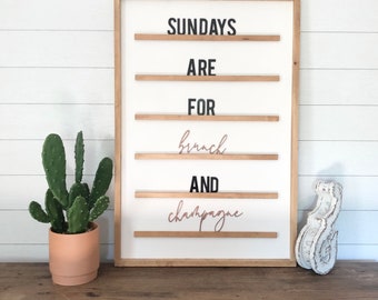 May include: A rectangular wall-mounted letter board with a white background and a light wood frame. The board features horizontal wooden slats holding black block letters and rose gold script letters that read: SUNDAYS ARE FOR brunch AND champagne.