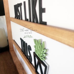 May include: Black wooden sign with the words "Just Like This" in large black letters. A small green plant is in a black pot in the lower right corner. The sign has a wooden frame and the text "Est. 2017 5th and Grace Home" is visible in the top center.