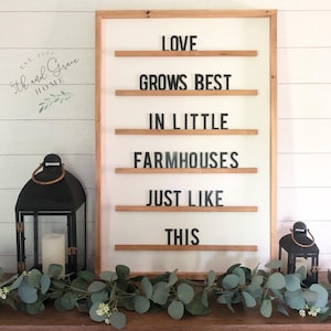 May include: A wooden framed sign with the text "LOVE GROWS BEST IN LITTLE FARMHOUSES JUST LIKE THIS" displayed on a white background. The sign is decorated with a garland of eucalyptus leaves.