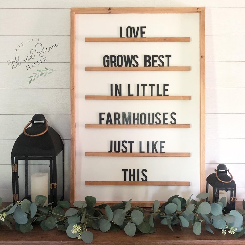 May include: A wooden framed sign with the text "LOVE GROWS BEST IN LITTLE FARMHOUSES JUST LIKE THIS" displayed on a white background. The sign is decorated with a garland of eucalyptus leaves.