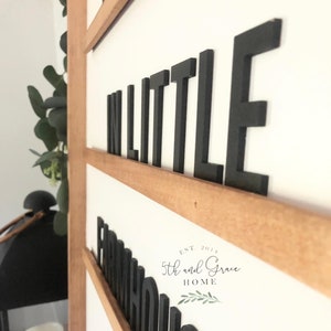 May include: A wooden sign with the words "In Little Farmhouse" in black block letters. The sign is on a white wall and has a brown wooden frame. The sign also includes the text "Est. 2014 5th and Grace Home" in black text.