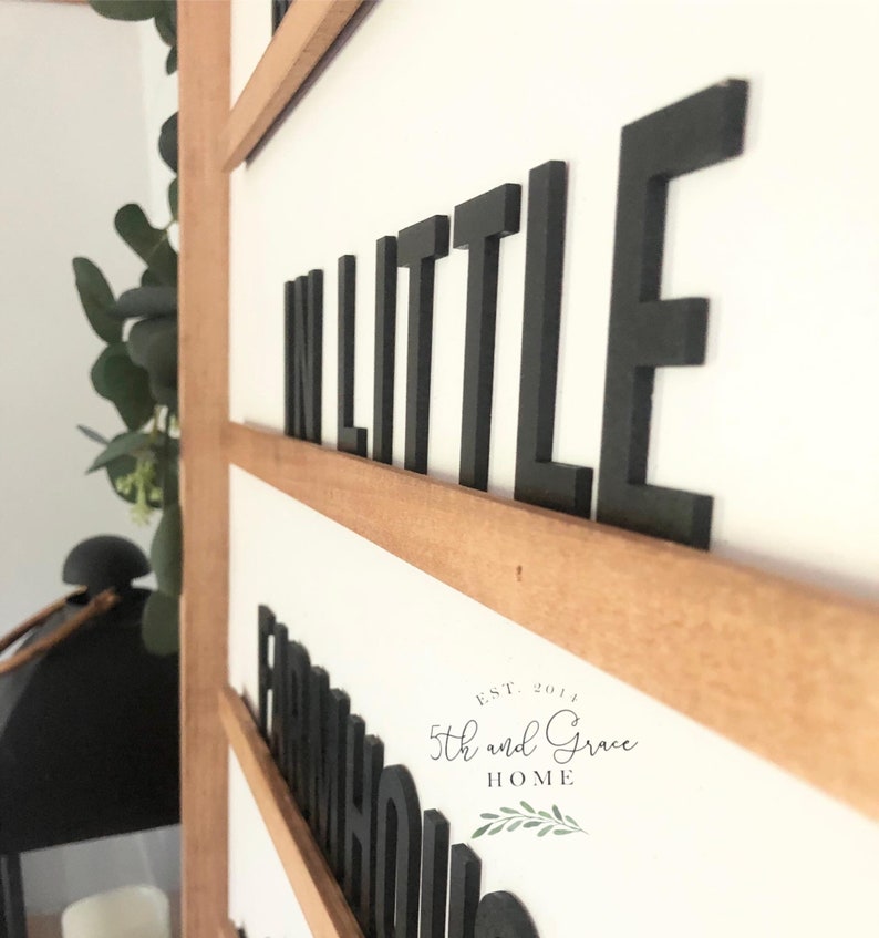 May include: A wooden sign with the words "In Little Farmhouse" in black block letters. The sign is on a white wall and has a brown wooden frame. The sign also includes the text "Est. 2014 5th and Grace Home" in black text.