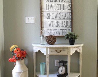 May include: A white painted wooden sign with the text "BE KIND, SPEAK TRUTH, LOVE OTHERS, SHOW GRACE, WORK HARD, BE GRATEFUL" hanging on a light green wall above a white painted half-moon table with a drawer and a shelf. The table has a vintage scale on the shelf and a glass vase with orange flowers on the floor.