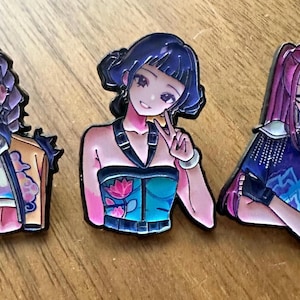 May include: Three anime-style enamel pins featuring female characters with different hairstyles and outfits. The pins have a glossy finish and are set against a wooden background. Each character has a unique pose and expression.