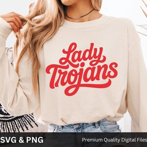 May include: Cream-colored long-sleeve sweatshirt with the words "Lady Trojans" in red script. The sweatshirt is made of a soft material and is suitable for casual wear. The image also includes the text "SVG & PNG" and "Premium Quality Digital Files".