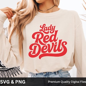 Puede incluir: Sudadera de manga larga color crema con las palabras "Lady Red Devils" en rojo, con una escritura estilizada. La sudadera se combina con unos vaqueros azules. La imagen también incluye el texto "SVG & PNG" y "Premium Quality Digital Files".