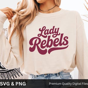May include: Cream-colored long-sleeve sweatshirt with the text "Lady Rebels" in a maroon, retro-style font. The sweatshirt is made of a soft material and is suitable for casual wear. The image also includes the text "SVG & PNG" and "Premium Quality Digital Files".