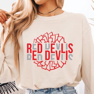 May include: A cream-colored long-sleeve sweatshirt featuring a graphic design with the text RED DEVILS in red and gray. The text is layered over a circular pattern of red geometric shapes, creating a bold, centered visual element on the chest.