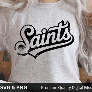 以下が含まれることがあります： 薄いグレーのスウェットシャツに、黒の擦り切れた筆記体で「Saints」の文字がプリントされています。テキストは黒で縁取られ、ヴィンテージ感があります。画像の下部には「SVG & PNG」と「Premium Quality Digital Files」の文字が表示されています。