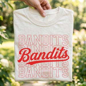 May include: A cream-colored t-shirt featuring the word BANDITS printed in a red, block-style varsity font across four horizontal lines, with the word Bandits written in a red, cursive script font layered over the center of the text.