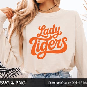 May include: Cream-colored long-sleeve sweatshirt with the words "Lady Tigers" in orange script. The text is in a retro font. The sweatshirt is made of a soft material. The bottom of the image shows the words "SVG & PNG" and "Premium Quality Digital Files".