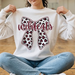 May include: White sweatshirt featuring a large bow design with a leopard print pattern in black and burgundy. The word "Wildcats" is written across the bow in burgundy script. The sweatshirt is a casual style.