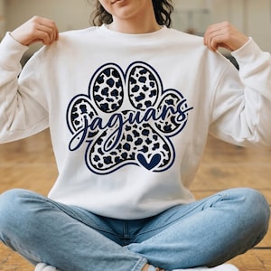 Op de afbeelding: Witte sweatshirt met een marineblauwe pootafdruk en een luipaardprint. Het woord "Jaguars" is in een cursief lettertype over de pootafdruk geschreven. Een klein marineblauw hart bevindt zich aan de onderkant van de poot.