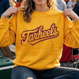 May include: A mustard yellow crewneck sweatshirt featuring the word Tarheels printed in a dark maroon, distressed script font across the chest. The garment has long sleeves and a ribbed neckline, providing a classic fit for casual wear.