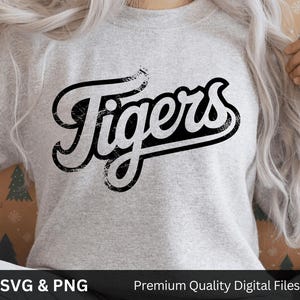 May include: A light gray sweatshirt with the word "Tigers" in a black, distressed, script font. The sweatshirt has long sleeves. The bottom of the image has the text "SVG & PNG" and "Premium Quality Digital Files".