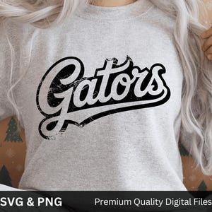 May include: A light gray sweatshirt with the word "Gators" in distressed black lettering. The text is in a script font with a black outline. The sweatshirt is a crew neck style.