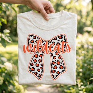 May include: Cream-colored t-shirt featuring a large bow design with a leopard print pattern in black and orange. The word "Wildcats" is written across the bow in orange script. The shirt is held up by a hand.