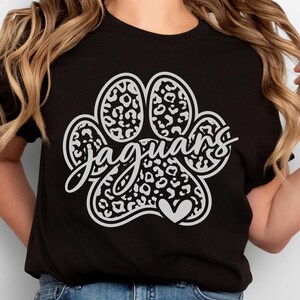 Op de afbeelding: Een zwart T-shirt met korte mouwen en een grote witte pootafdruk met een luipaardpatroon. Het woord "jaguars" staat in een wit cursief lettertype in het midden van de poot, met een klein wit hartje aan de onderkant van de afbeelding.