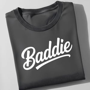 May include: A folded charcoal gray t-shirt with the word "Baddie" in white script across the front. The shirt has a crew neck and a simple, clean design, suitable for casual wear.