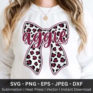 May include: White sweatshirt with a large bow design. The bow has a leopard print pattern in black and maroon, with the word "aggie" in maroon script. The image includes the text "SVG - PNG - EPS - JPEG - DXXF" at the bottom.