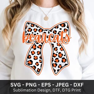 May include: White sweatshirt featuring a large bow design with a leopard print pattern in black and orange. The word "Bearcats" is written in orange script across the bow. The bottom of the image includes text: SVG - PNG - EPS - JPEG - DXF.