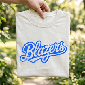 Peut inclure: T-shirt crème avec le mot "Blazers" en script bleu vieilli sur le devant. Le t-shirt est présenté sur un fond vert flou, suggérant un cadre extérieur. Le texte a un aspect vintage et usé.