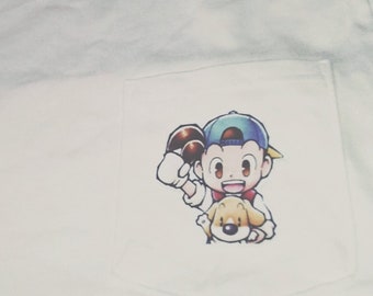 Könnte beinhalten: Weißes T-Shirt mit einer Tasche, die eine Cartoon-Illustration eines Jungen mit einer blauen Mütze und einem Hund zeigt. Der Junge hält einen roten und weißen Ball.