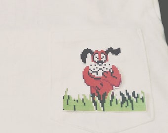 May include: White t-shirt with a pixelated image of a red dog in a pocket. The dog is holding a bone and is surrounded by green grass.