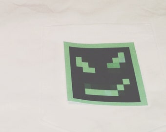 Könnte beinhalten: Weißes T-Shirt mit einem grünen und schwarzen pixelartigen Gesicht auf der Tasche.