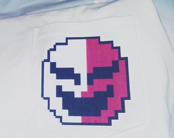 Könnte beinhalten: Weißes T-Shirt mit einem pixeligen Gesichtsdesign auf der Tasche. Das Gesicht ist vertikal geteilt, eine Hälfte in Weiß und die andere in Rosa. Der Mund ist dunkelblau.