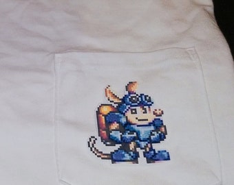 Op de afbeelding: Wit T-shirt met een blauw en geel gepixelde figuur op de zak. Het figuur draagt een helm en een bril.