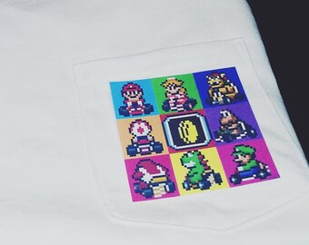 May include: White t-shirt with a pocket featuring a colorful pixelated graphic of characters from a racing video game. The design includes Mario, Luigi, and other familiar figures in their karts.