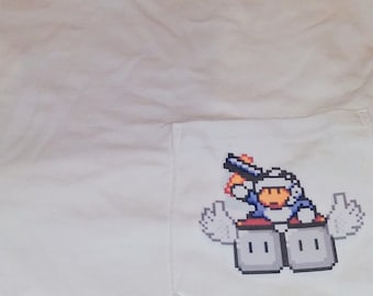 May include: White t-shirt with a pocket featuring a pixelated character holding a gun and standing on two blocks. The character is wearing a blue and white suit and has a yellow face.
