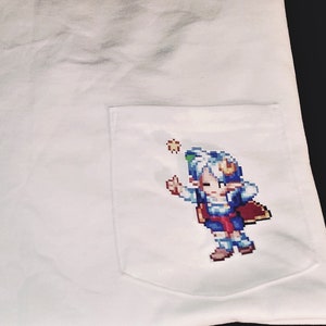 May include: White t-shirt with a pocket featuring a pixelated character design. The character is blue, red, and white and is holding a star.