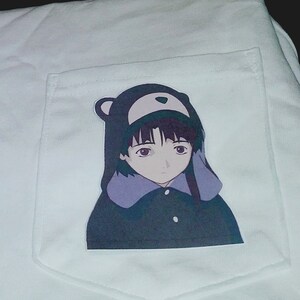 May include: White t-shirt with a pocket featuring a printed image of an anime character wearing a hooded jacket with bear ears.