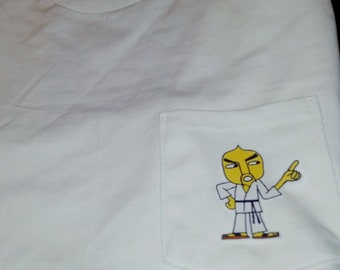 Op de afbeelding: Wit T-shirt met een zak met daarop een cartoonfiguur dat een wit karatepak draagt met een zwarte band en naar rechts wijst. Het figuur heeft een geel gezicht met zwarte ogen en wenkbrauwen, een zwarte neus en een rode mond.