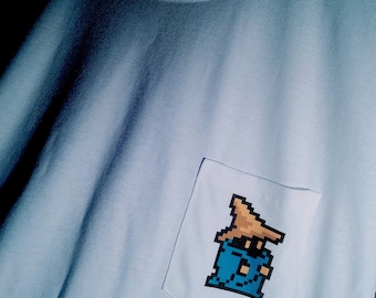 Op de afbeelding: Een lichtblauw t-shirt met een klein, gepixeld beeld van een tovenaar met een bruine hoed op de zak.