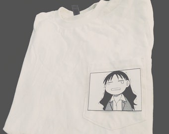 Könnte beinhalten: Weißes T-Shirt mit einer Tasche, die eine schwarz-weiße Manga-Illustration einer Person mit langen dunklen Haaren und neutralem Gesichtsausdruck zeigt. Das T-Shirt ist gefaltet und aus weichem Stoff.